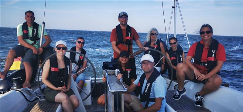 Eine Gruppe von Menschen sitzt auf einem Segelboot im Cockpit und lächelt in die Kamera. Im Hintergrund das blaue Wasser.