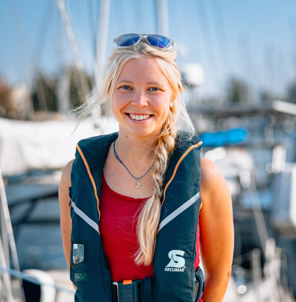 Eine Person mit blonden Haaren & geflochtenem Zopf und Sonnenbrille auf dem Kopf lächelt in die Kamera. Im Hintergrund sind verschwommen die Masten von Segelbooten zu sehen.