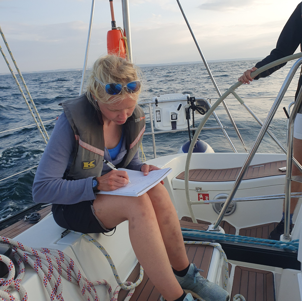 Eine Person sitzt auf einem Segelboot und schreibt auf einen Zettel mit einem Stift. Im Hintergrund sieht man Wasser.