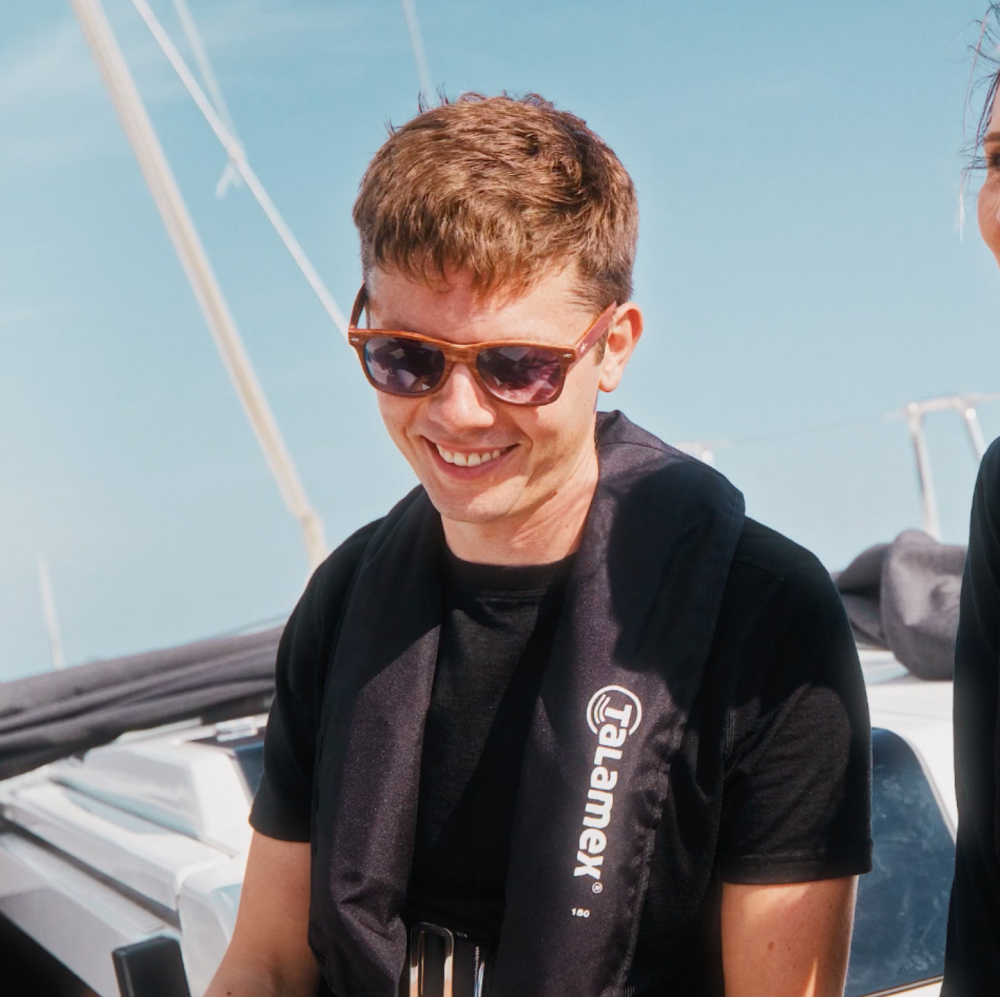 Ein junger Mann mit Sonnenbrille lacht und schaut auf den Boden. Im Hintergrund der blaue Himmel.