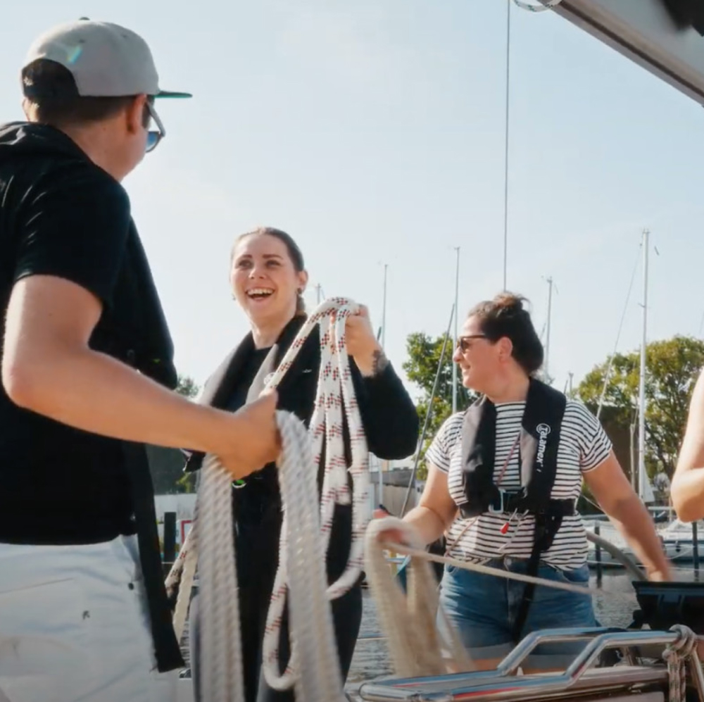 Ein Team hantiert auf einem Segelboot mit Leinen und lacht sich unter einander an.