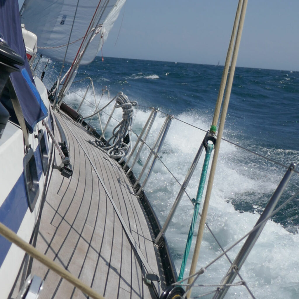 Blick entlang des Seitendecks auf einem Segelboot nach Vorn. Ein Vorsegel ist zu sehen und das Wasser spritzt an der Bordwand.