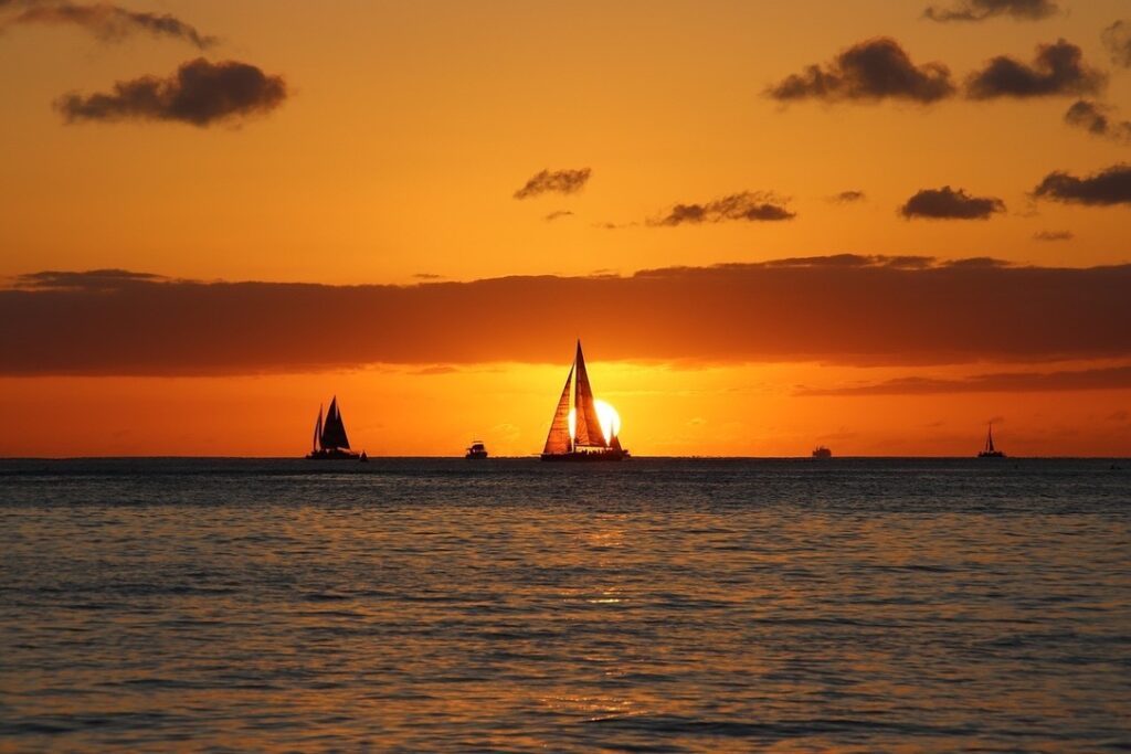 Blick in einen Sonnenuntergang auf See. Die Sonne ist nah am Horizont, alles ist orange und golden. Im Vordergrund ruhige See und vor der Sonne ein Segelboot und weitere am Horizont.