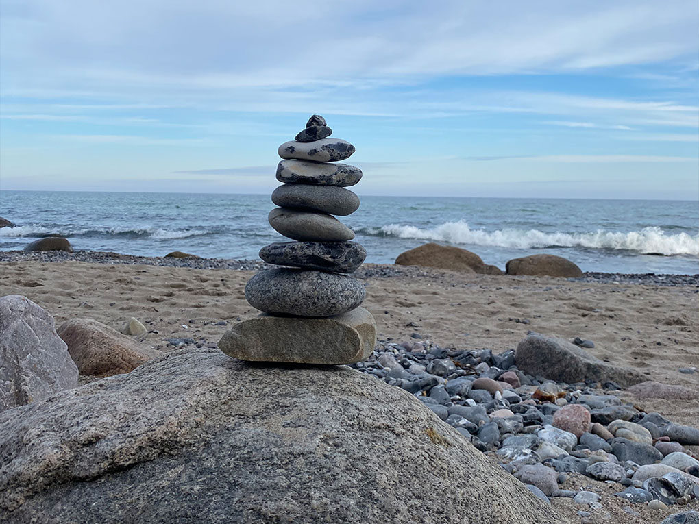 Ein kleiner Steinhaufen steht aufgetürmt auf einem großen Stein. Im Hintergrund der Strand und das Meer.