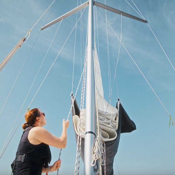 Eine Person steht vor einem Segelmast und blickt an ihm entlang nach oben, während sie eine Leine in der Hand hält und sich das Segel nach oben bewegt.