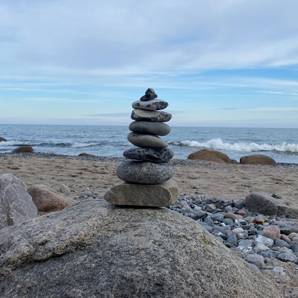 Ein kleiner Steinhaufen steht aufgetürmt auf einem großen Stein. Im Hintergrund der Strand und das Meer.